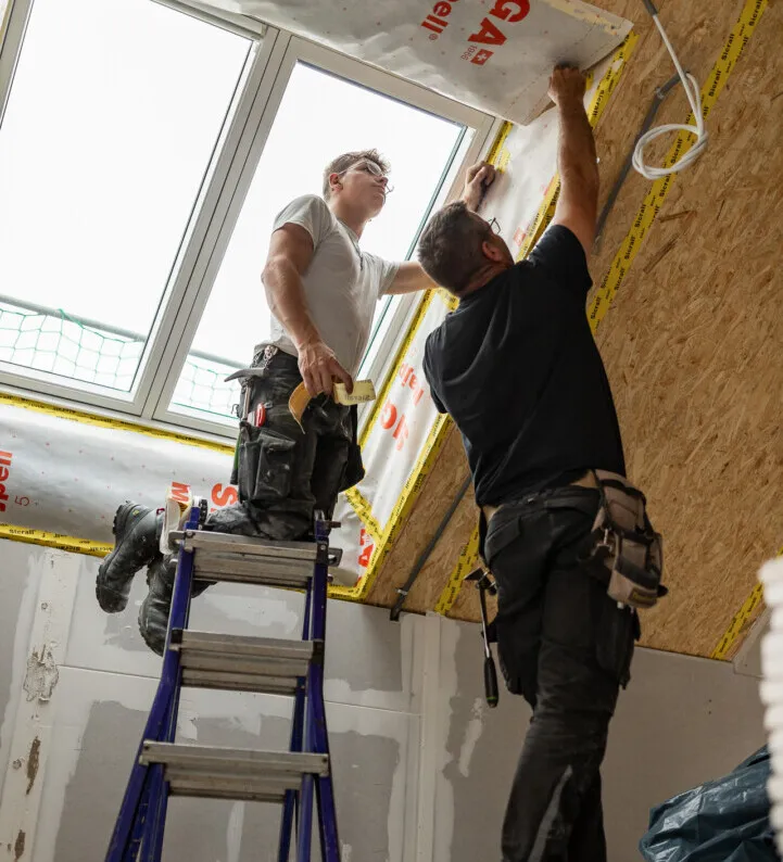 Zwei Männer arbeiten an einem Dach in einem unvollendeten Dachboden. Werkzeuge und Materialien sind sichtbar.